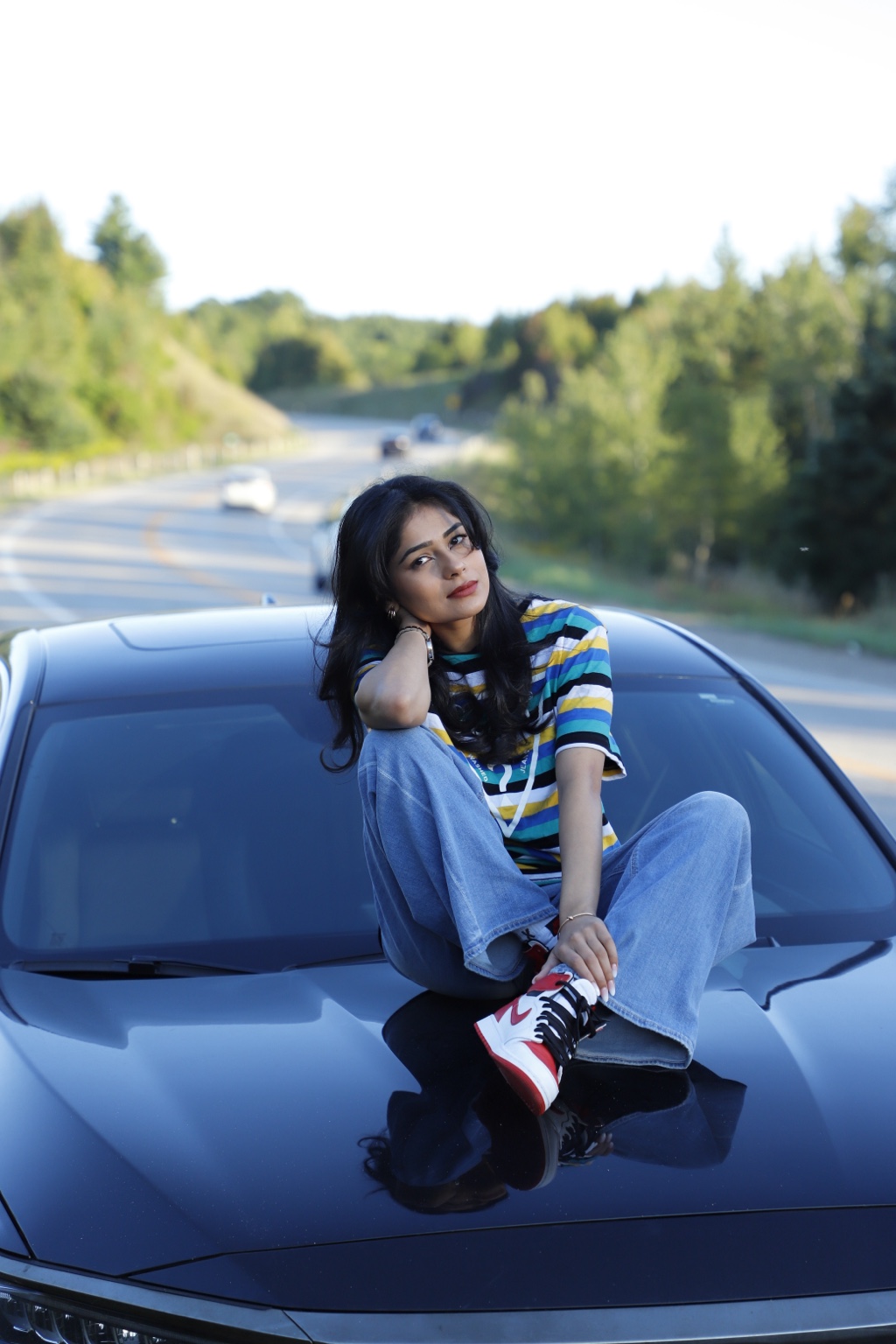 Komal Sandhu in a striped top and jeans seated on a car hood