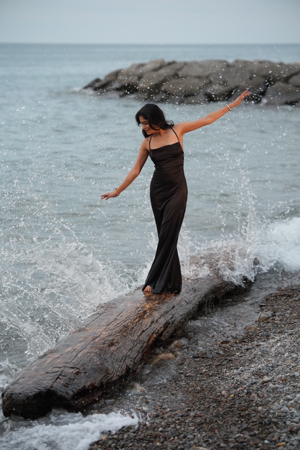 Komal Sandhu in a black dress balancing on a driftwood log with waves splashing around her